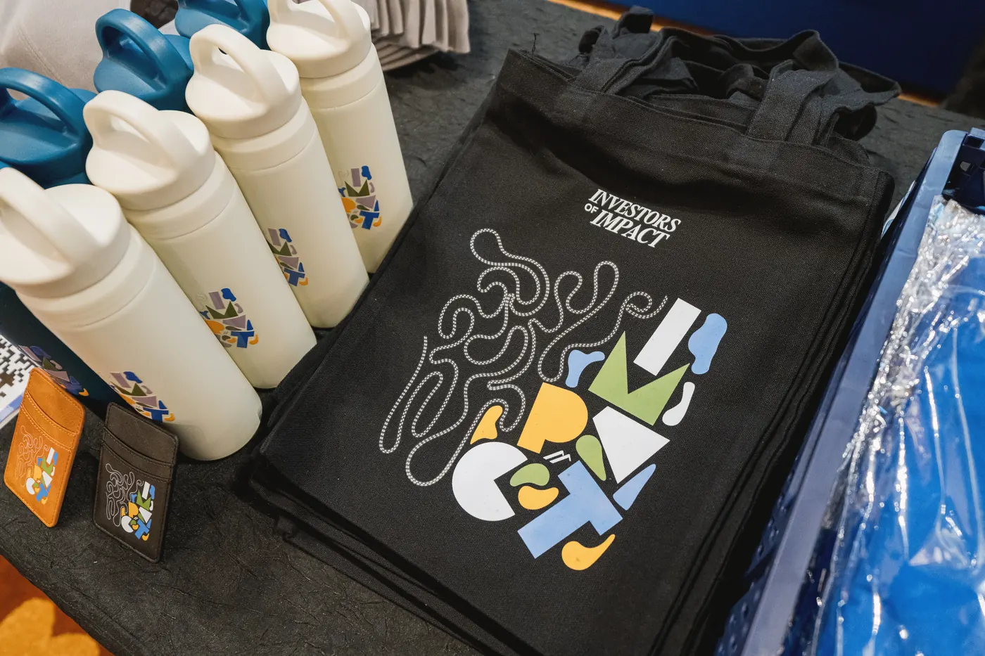 A black tote bag printed with the IMPACT artwork, beside branded bottles